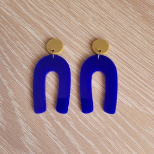 The Blue Wishbone earrings start with an olive green solid acrylic circle stud, and then continue to an upside-down U shape in an opaque blue acrylic. The earrings are sitting on a wooden table.