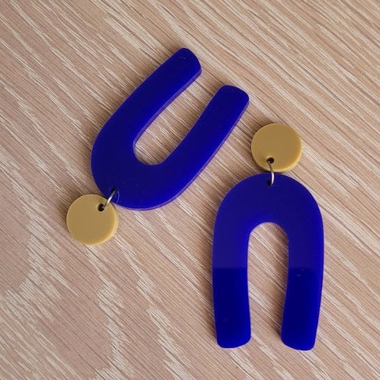 The Blue Wishbone earrings start with an olive green solid acrylic circle stud, and then continue to an upside-down U shape in an opaque blue acrylic. The earrings are sitting on a wooden table.