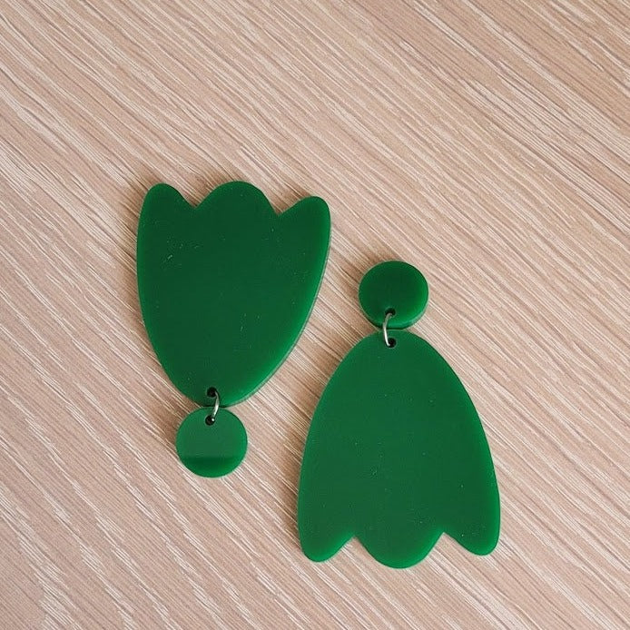 The Green Tulip Earrings begin with a solid round green acrylic stud, and continue to a solid green acrylic tulip shape. The earrings are sitting on a wooden table and are pictured with one earring upside-down.