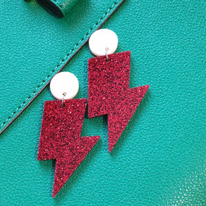Red glitter lightning bolt drop earrings, with a light pink round stud.  The earrings are made from acrylic, and are resting on a background of green leather.