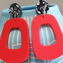 Load image into Gallery viewer, The Lola earrings have a black and silver chunky glitter round stud and then drop down to an orangey red rounded oblong shape.  They are hanging from a blue clay vase.  This is a close up image.