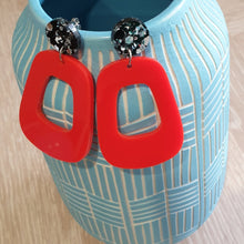 Load image into Gallery viewer, The Lola earrings have a black and silver chunky glitter round stud and then drop down to an orangey red rounded oblong shape. They are hanging from a blue clay vase.  This is a birds-eye view.