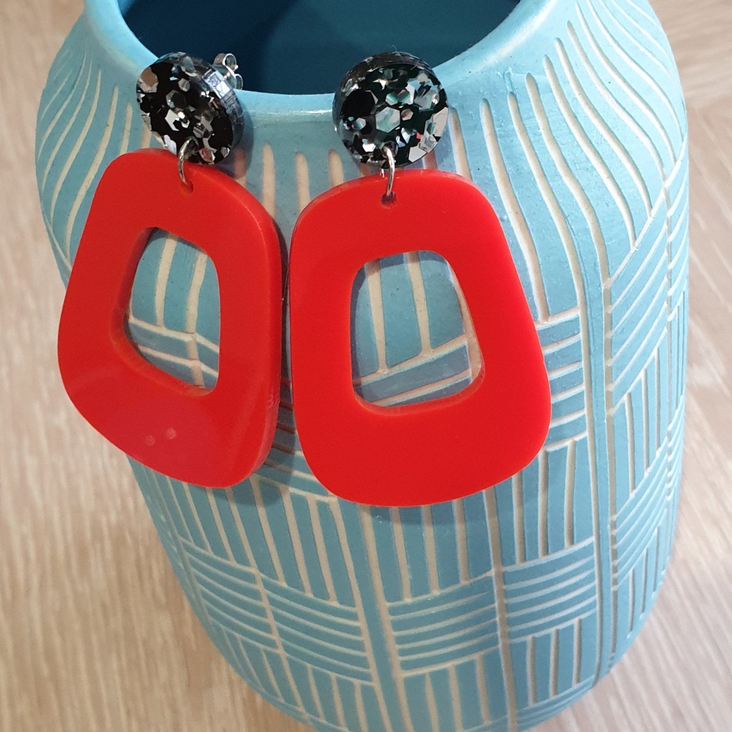 The Lola earrings have a black and silver chunky glitter round stud and then drop down to an orangey red rounded oblong shape. They are hanging from a blue clay vase.  This is a birds-eye view.