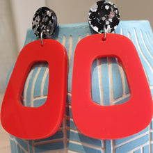 Load image into Gallery viewer, The Lola earrings have a black and silver chunky glitter round stud and then drop down to an orangey red rounded oblong shape. They are hanging from a blue clay vase.