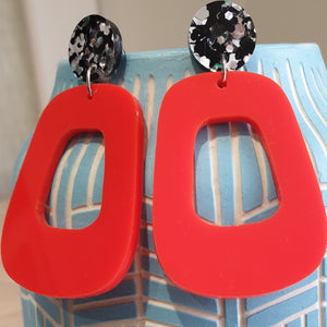 The Lola earrings have a black and silver chunky glitter round stud and then drop down to an orangey red rounded oblong shape. They are hanging from a blue clay vase.