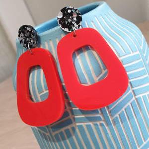 The Lola earrings have a black and silver chunky glitter round stud and then drop down to an orangey red rounded oblong shape. They are hanging from a blue clay vase.  This is a zoomed out view.