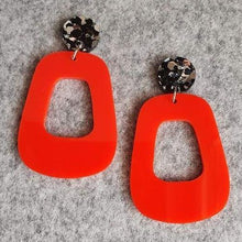 Load image into Gallery viewer, The Lola earrings have a black and silver chunky glitter round stud and then drop down to an orangey red rounded oblong shape. They are laying on a background of light grey felt.