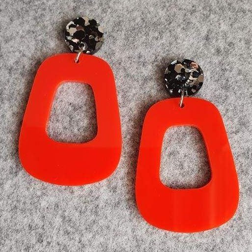 The Lola earrings have a black and silver chunky glitter round stud and then drop down to an orangey red rounded oblong shape. They are laying on a background of light grey felt.