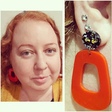 Load image into Gallery viewer, The Lola earrings have a black and silver chunky glitter round stud and then drop down to an orangey red rounded oblong shape. This image shows the Lola earrings on a woman.