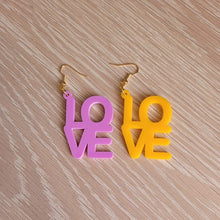 Load image into Gallery viewer, The Love Earrings consist of one purple earring and one amber earring, both in the shape of the word &quot;Love&quot;.  They are sitting on a wooden table.