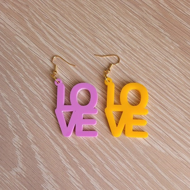 The Love Earrings consist of one purple earring and one amber earring, both in the shape of the word "Love".  They are sitting on a wooden table.