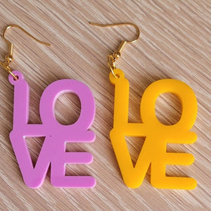 The Love Earrings consist of one purple earring and one amber earring, both in the shape of the word "Love". They are sitting on a wooden table.  This is a close up image.