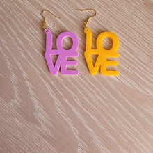 Load image into Gallery viewer, The Love Earrings consist of one purple earring and one amber earring, both in the shape of the word &quot;Love&quot;. They are sitting on a wooden table.