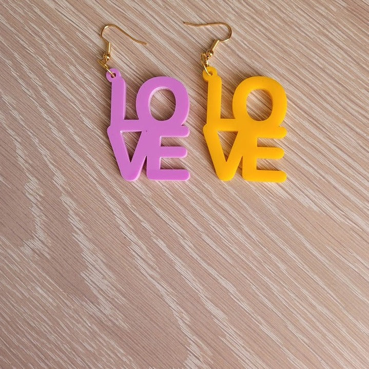 The Love Earrings consist of one purple earring and one amber earring, both in the shape of the word "Love". They are sitting on a wooden table.