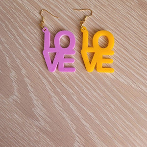The Love Earrings consist of one purple earring and one amber earring, both in the shape of the word "Love". They are sitting on a wooden table.