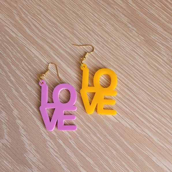 The Love Earrings consist of one purple earring and one amber earring, both in the shape of the word "Love". They are sitting on a wooden table.