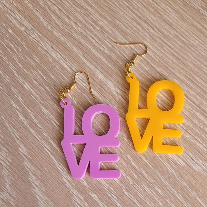 The Love Earrings consist of one purple earring and one amber earring, both in the shape of the word "Love". They are sitting on a wooden table.