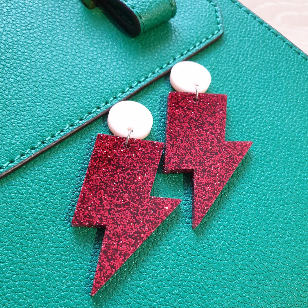 Red glitter lightning bolt drop earrings, with a light pink round stud.  The earrings are made from acrylic, and are resting on a green leather bag.