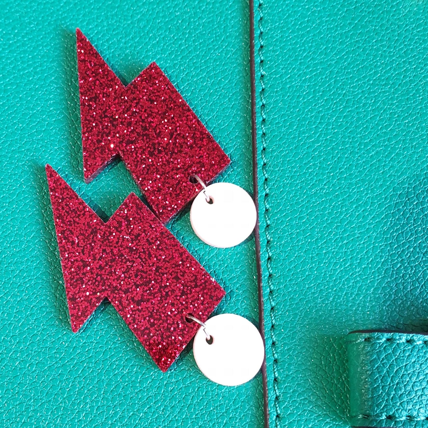 Red glitter lightning bolt drop earrings, with a light pink round stud.  The earrings are made from acrylic, and are resting on a green leather bag.  The earrings are positioned diagonally, and upside down.