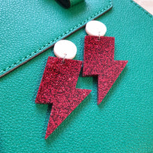 Load image into Gallery viewer, Red glitter lightning bolt drop earrings, with a light pink round stud.  The earrings are made from acrylic, and are resting on a green leather bag.  The earrings are positioned diagonally.