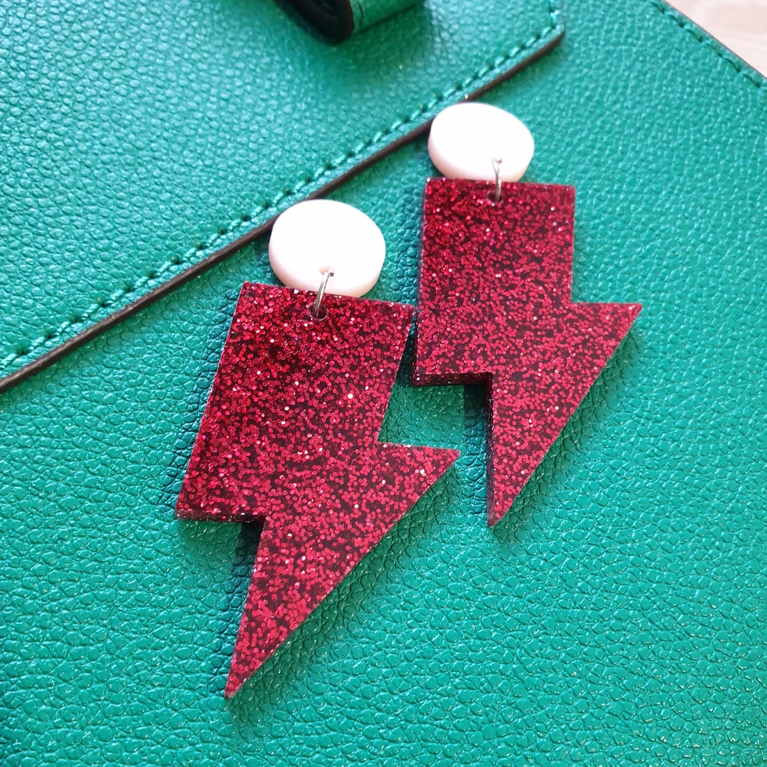 Red glitter lightning bolt drop earrings, with a light pink round stud.  The earrings are made from acrylic, and are resting on a green leather bag.  The earrings are positioned diagonally.