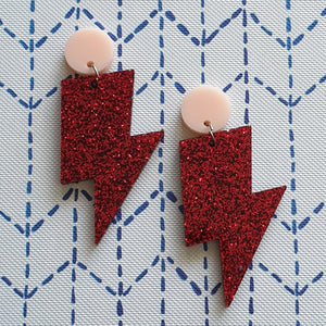 Red glitter lightning bolt drop earrings, with a light pink round stud.  The earrings are made from acrylic, and are resting on a background of blue and white dots.